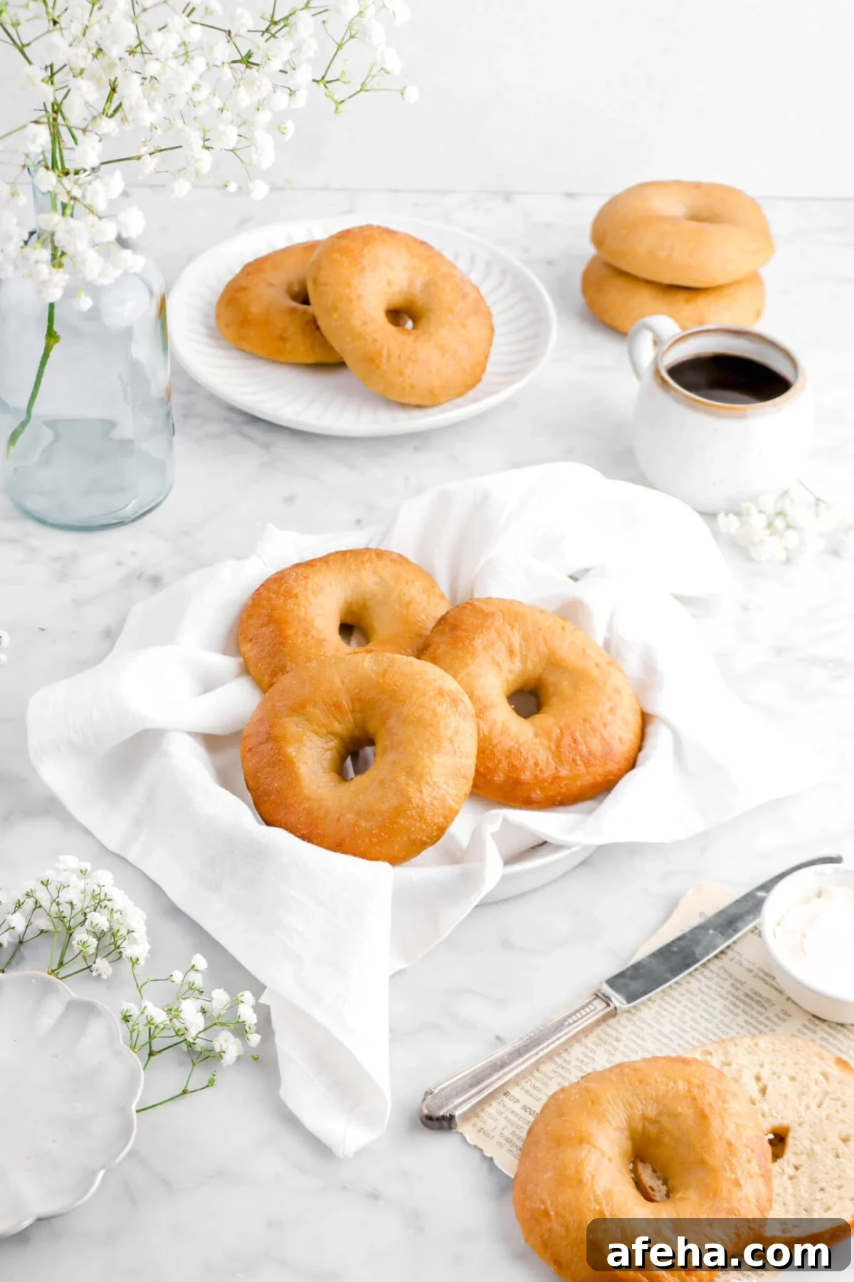 angled shot of three bagels in pie plate with white napkin, four bagels behind with a glass vase of flowers, and cup of coffee