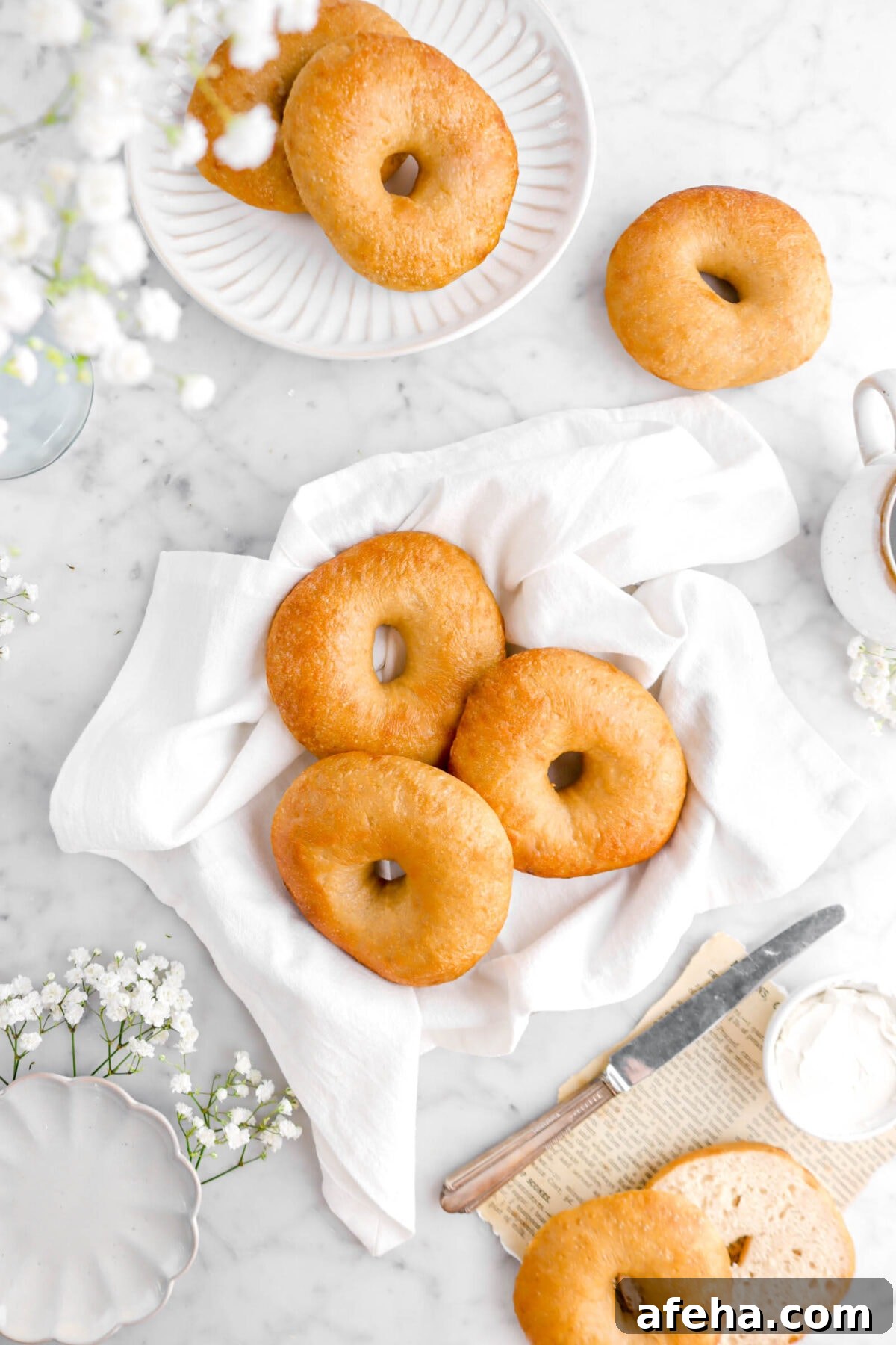 overhead shot of three bagels in white napkin with two bagels on a plate above, another bagel on marble counter, and one cut open on book page with knife and bowl of cream cheese beside