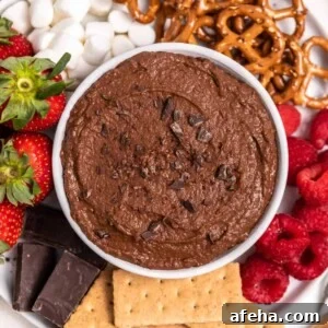 White bowl with chocolate brownie batter hummus on plate with berries, graham crackers and other dippers.