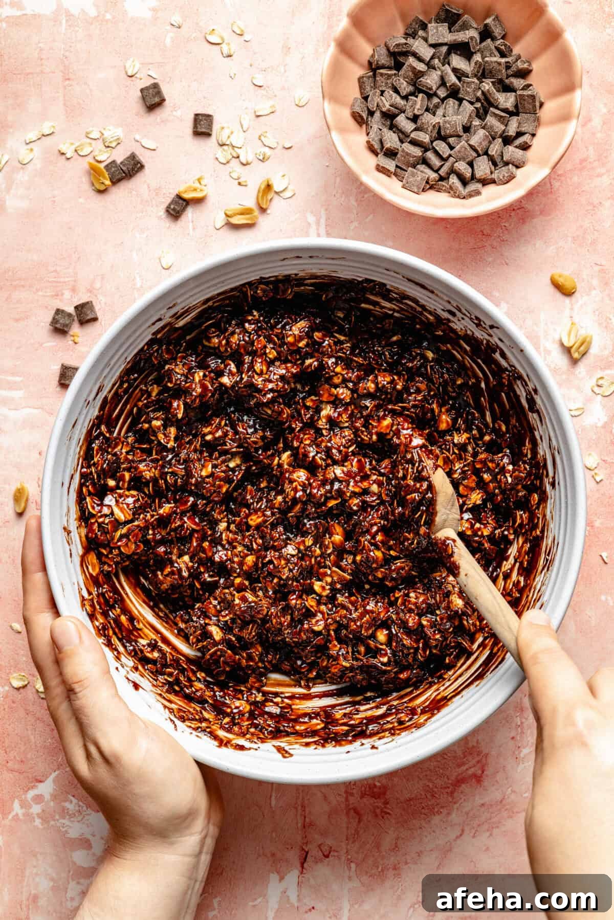 a mixing bowl with chocolate, oats, and salted peanuts on a light pink background