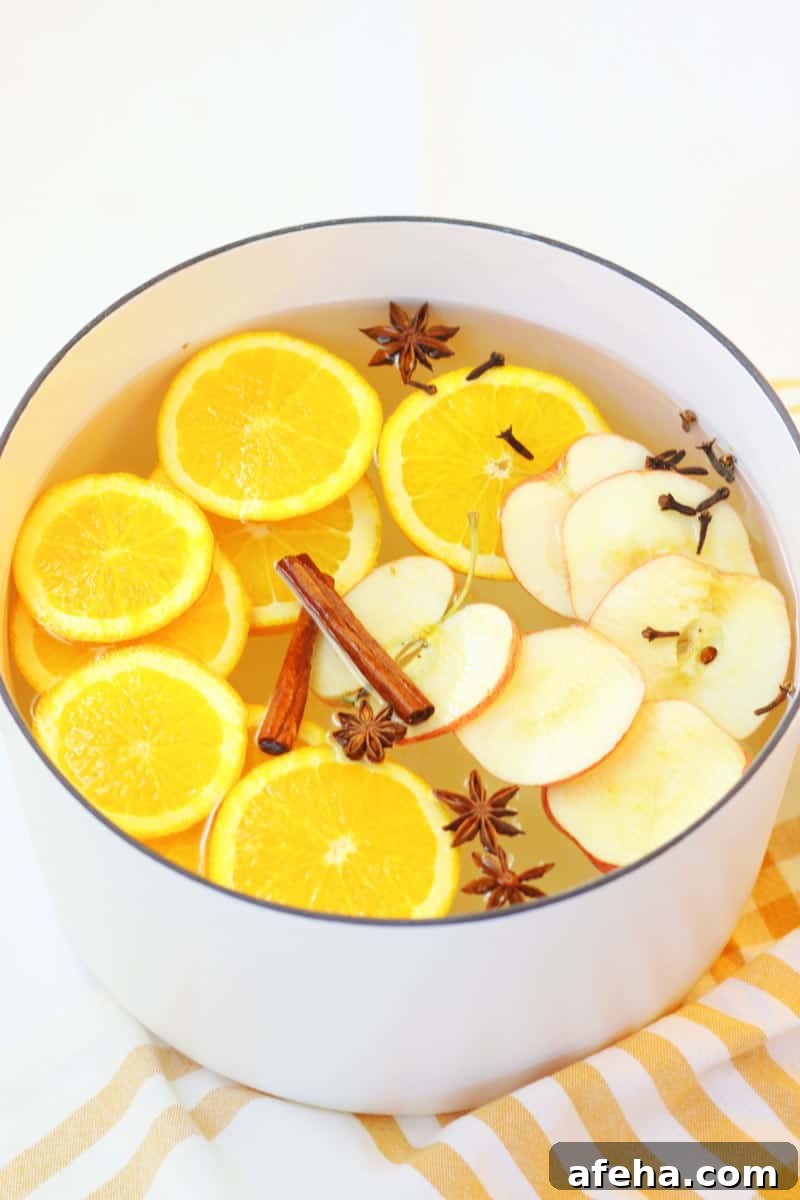 a white dutch oven with a striped yellow and white towel under it. It is filled with water and a fresh homemade potpourri made of oranges, apples and spices.