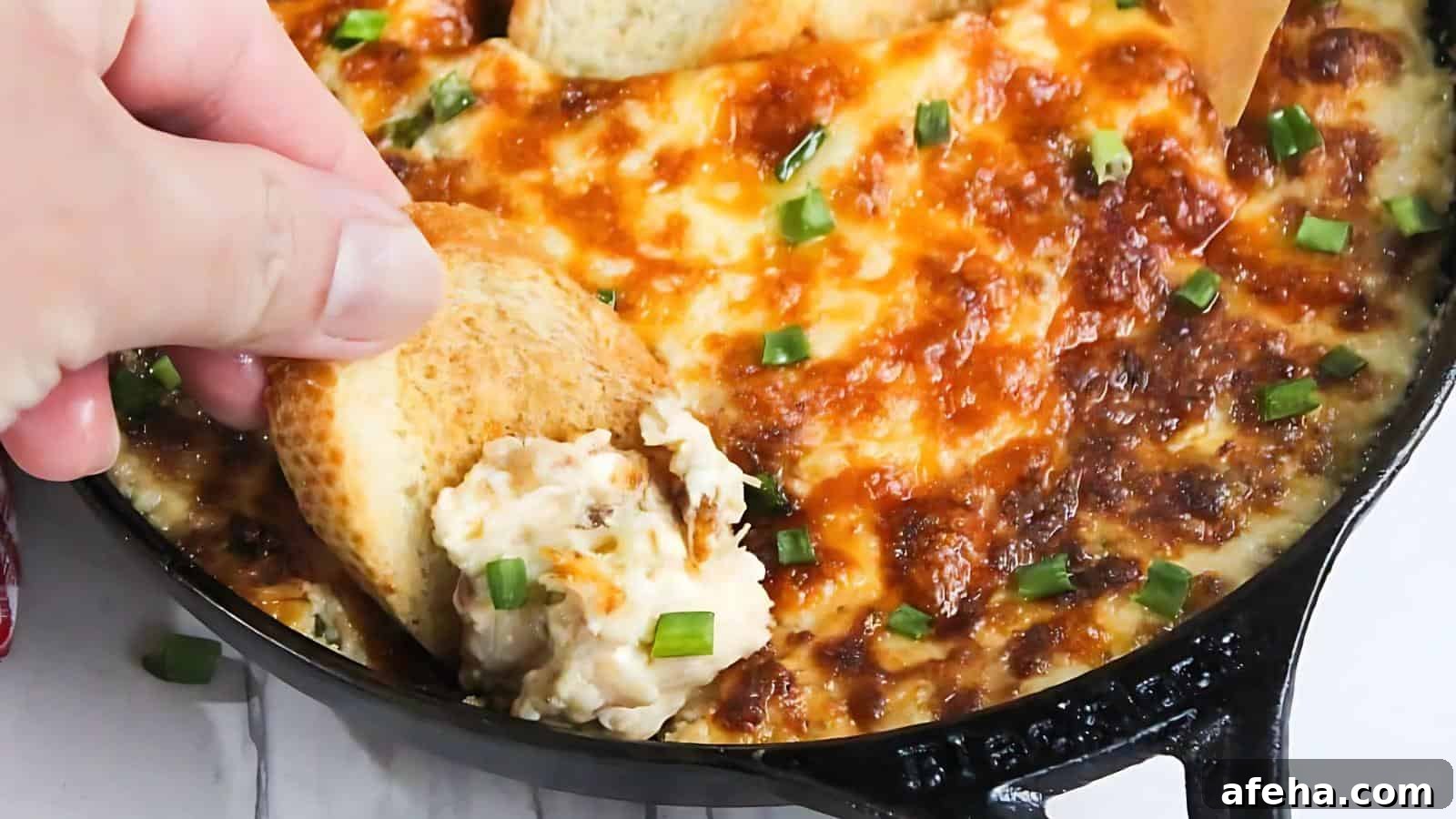 Close-up of a cheesy baked dip topped with chopped green onions, with a hand dipping a piece of toasted bread into it, in a black skillet.