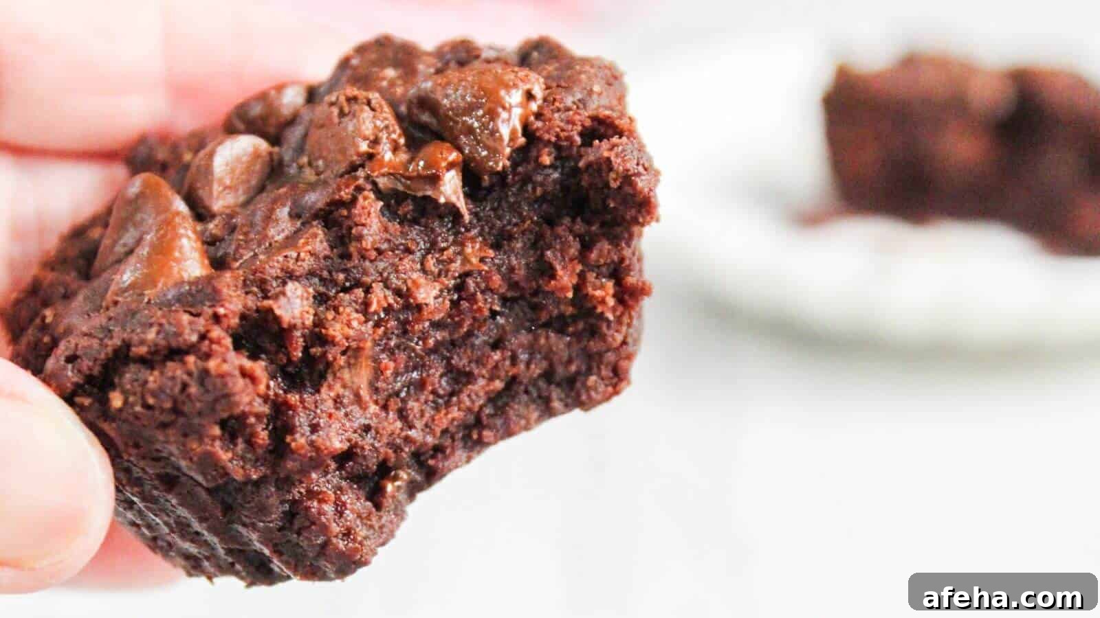 Close-up of a hand holding a half-eaten chocolate brownie with gooey chocolate chunks, with another partial brownie blurred in the background.