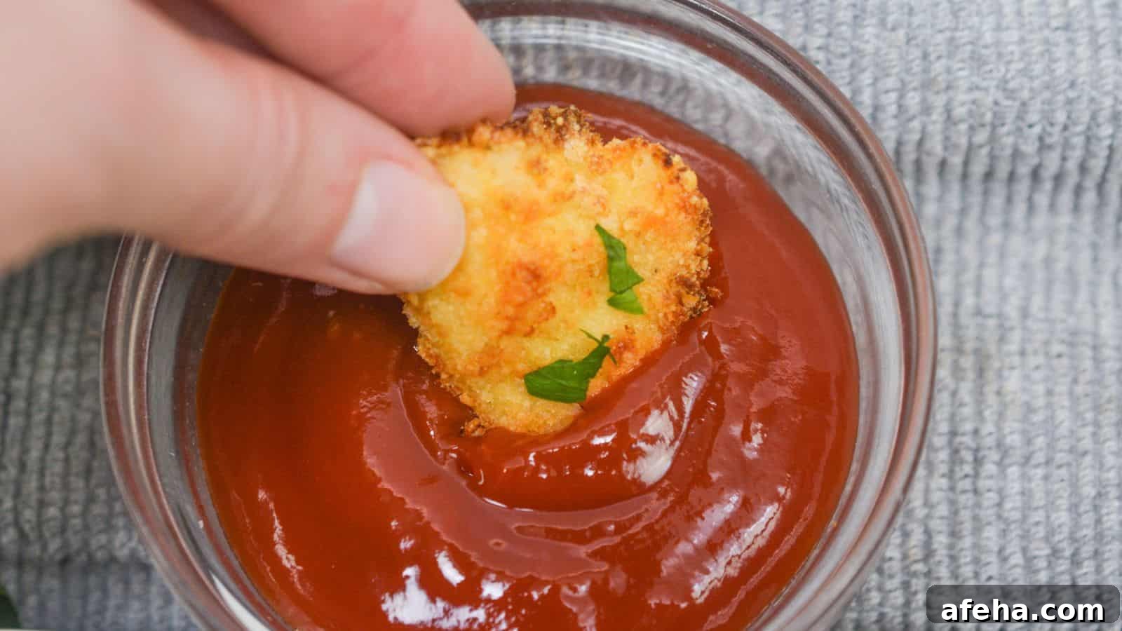 Hand dipping a chicken nugget into a bowl of ketchup, with a gray cloth background.
