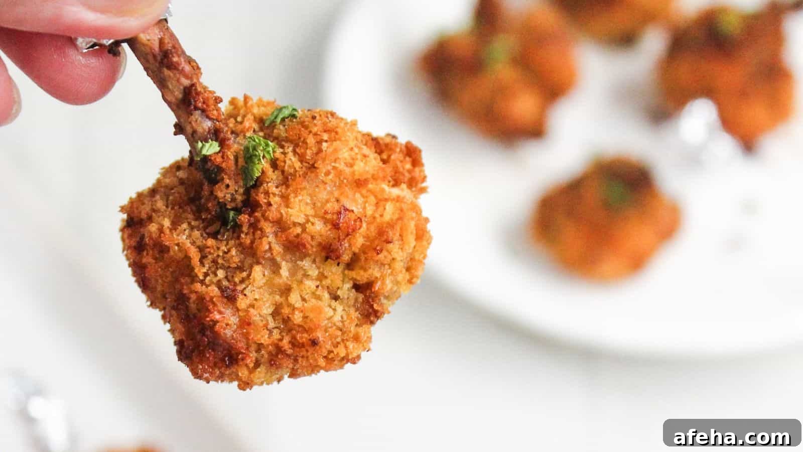 A close-up of a hand holding a crispy breaded chicken drumstick, with more drumsticks on a white plate in the background.