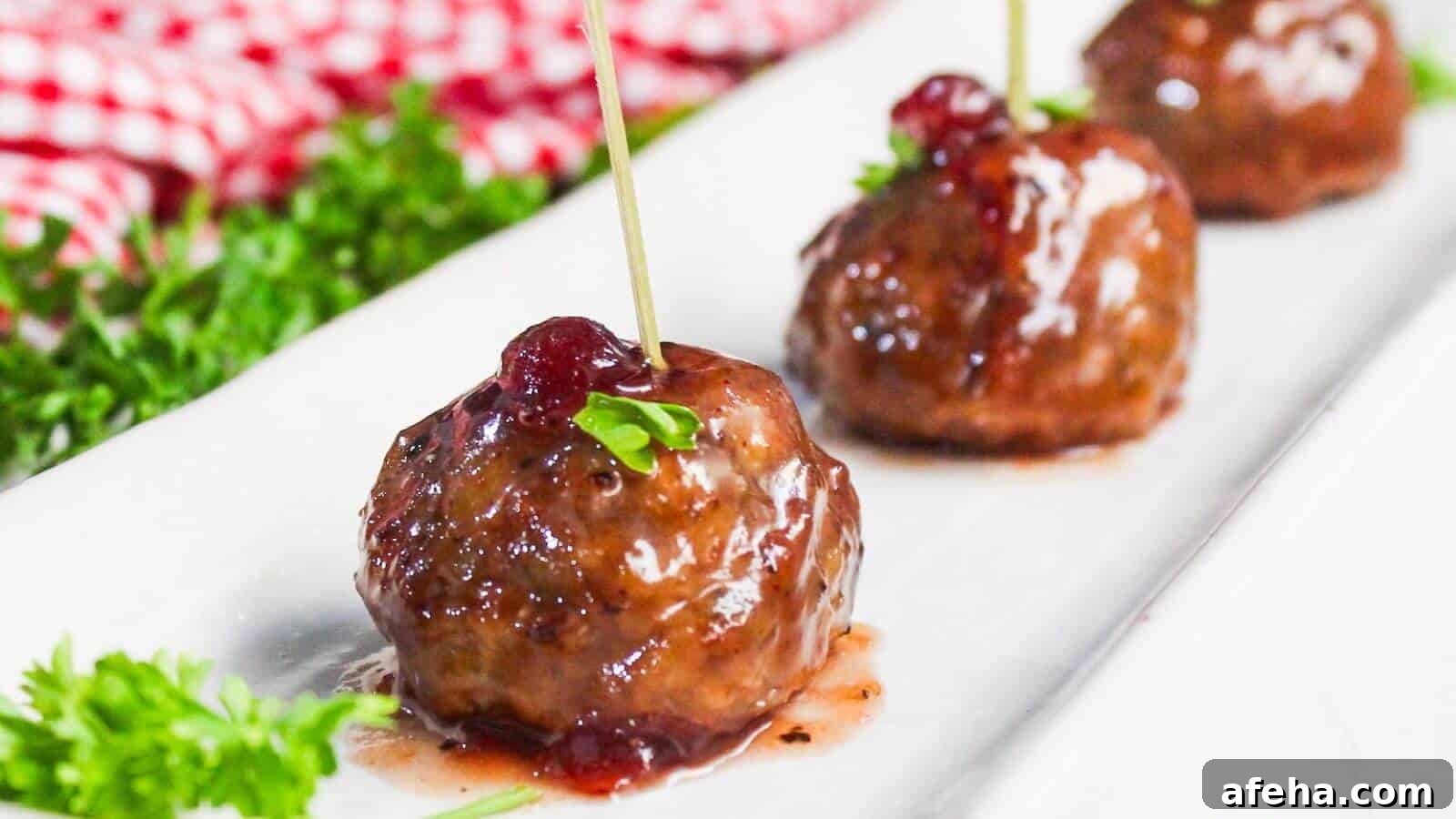 Three glazed meatballs are arranged in a row on a white plate, each garnished with a stick and a small sprig of parsley. A red checkered cloth and some fresh parsley are in the background.
