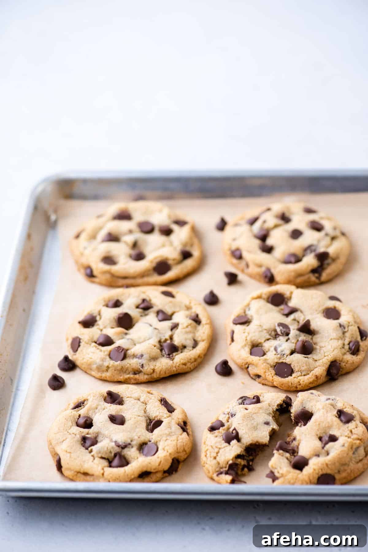 6 chocolate chip cookies on a parchment-lined baking sheet, with one broken in half