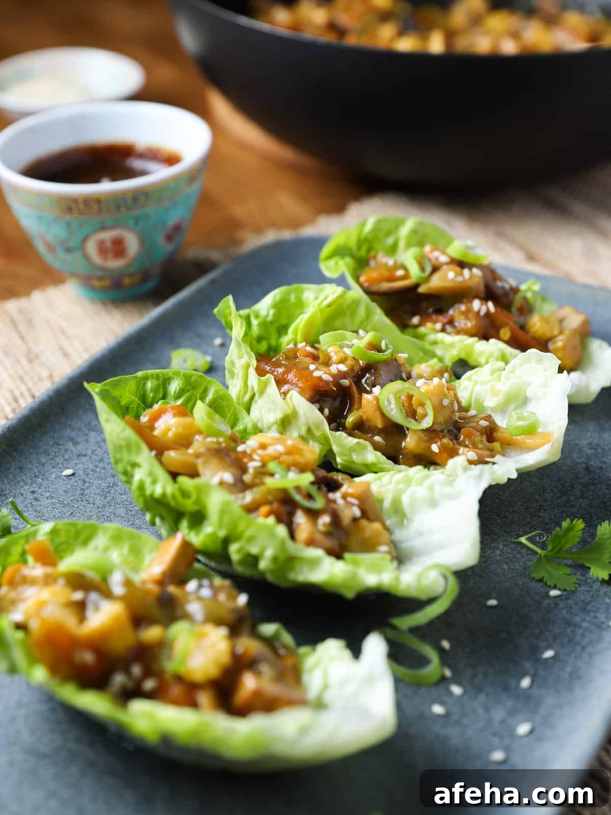 Vegan San Choy Bau Recipe: Flavorful Plant-Based Lettuce Wraps 2 Photo of 4 lettuce leaves with vegan san choy bau filling inside, placed on a blue-grey oblong plate with pan in the background and a chinese dish of hoisin sauce to the side.