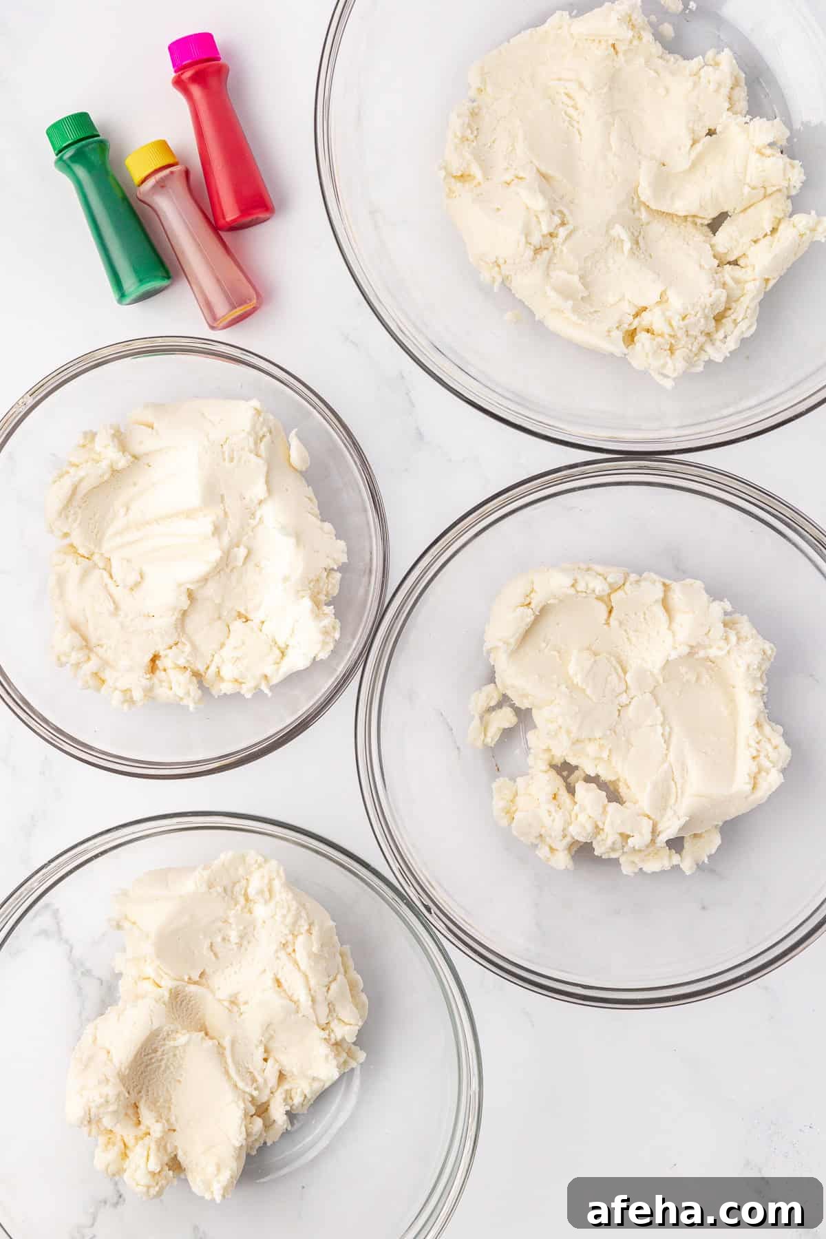 three bottles of food coloring (green, yellow, pink) beside four bowls of white butter mint dough