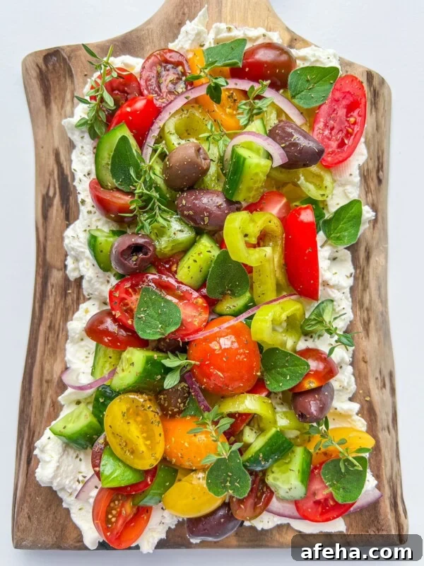 A vibrant Greek Salad Feta Board features cherry tomatoes, cucumbers, olives, red onion, peppers, and fresh herbs atop whipped feta, beautifully arranged on a rustic wooden serving board.