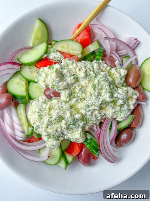 A bowl of Greek Feta Salad with sliced cucumbers, tomatoes, red onions, Kalamata olives, and a generous dollop of tzatziki sauce on top, with a spoon resting in the salad.