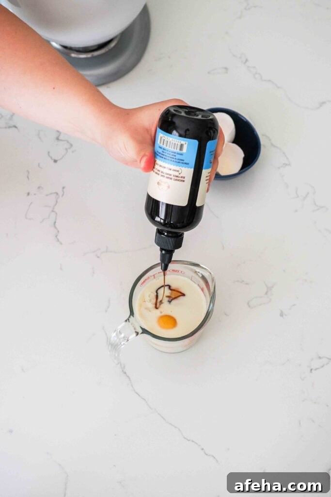 A hand squeezes vanilla bean paste into a measuring cup with milk and eggs.