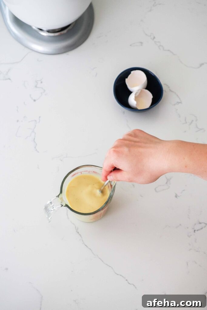A hand whisks eggs, milk, and vanilla bean paste together in a measuring cup.