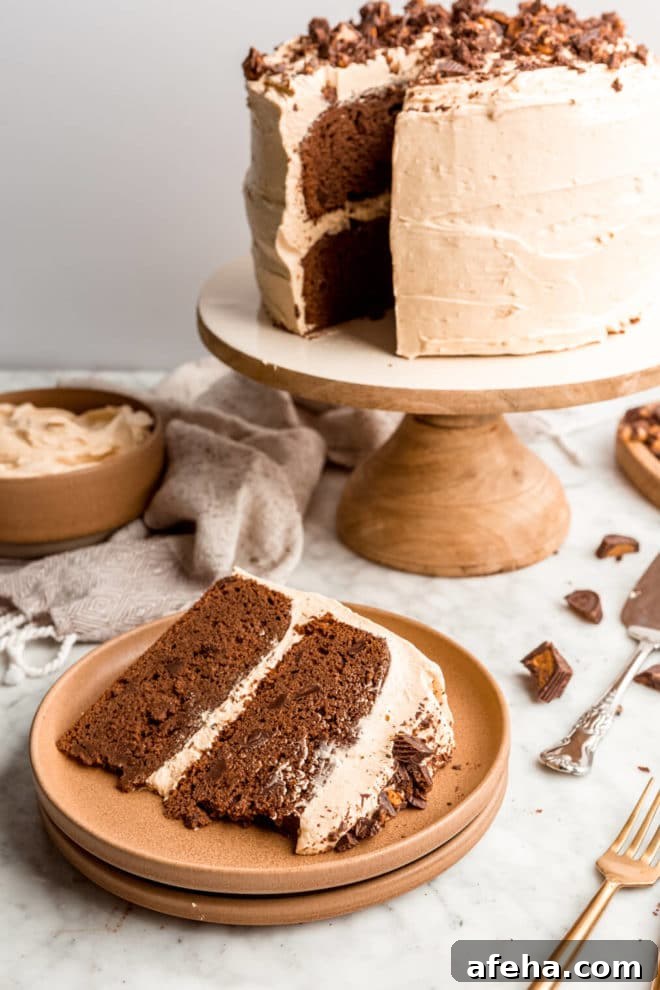 Slice of double layer chocolate cake on a clay-colored plate with a wooden cake stand in the background with a double layer chocolate cake coated in tan, peanut butter buttercream.