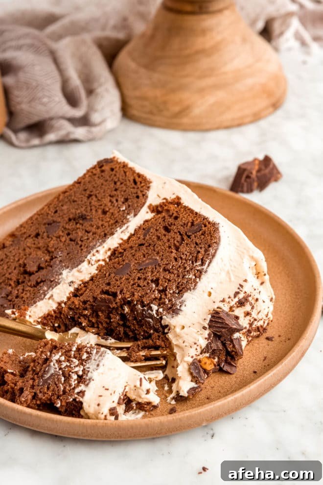 Slice of chocolate layer cake with peanut butter frosting on a brown plate with a bite of cake on a fork. There is a cake stand in the background.