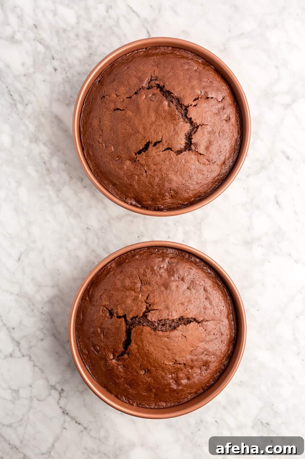 Two baked chocolate cakes in round cake pans on a grey and white marble surface.