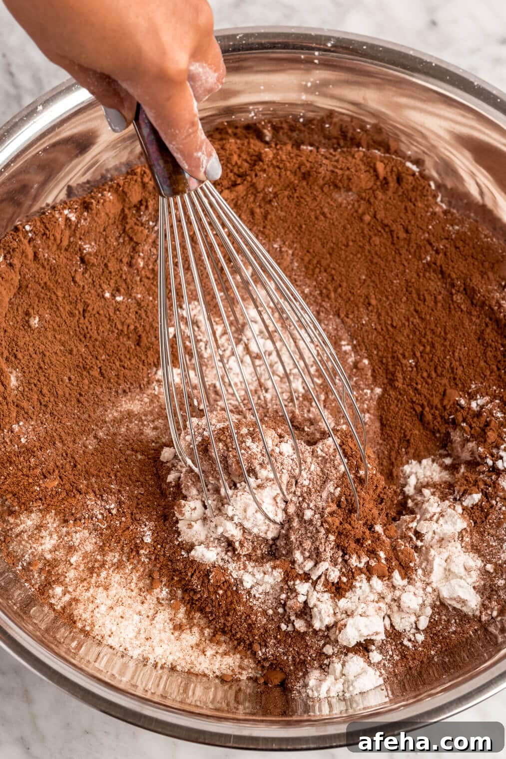 Dry chocolate cake ingredients being whisked together in a large, metal bowl.