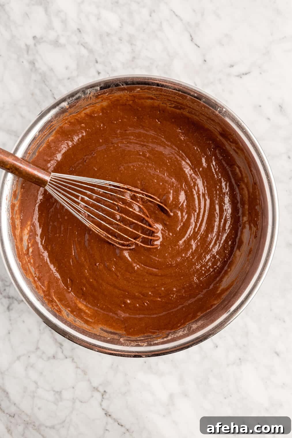 Chocolate cake batter whisked until smooth in a large metal bowl on a grey and white marble surface.