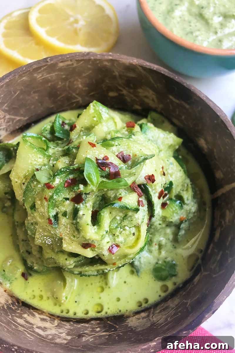 Vertical image of thinly sliced cooked vegetables covered in a vibrant green sauce drizzled with olive oil in a wooden bowl next to lemon slices.