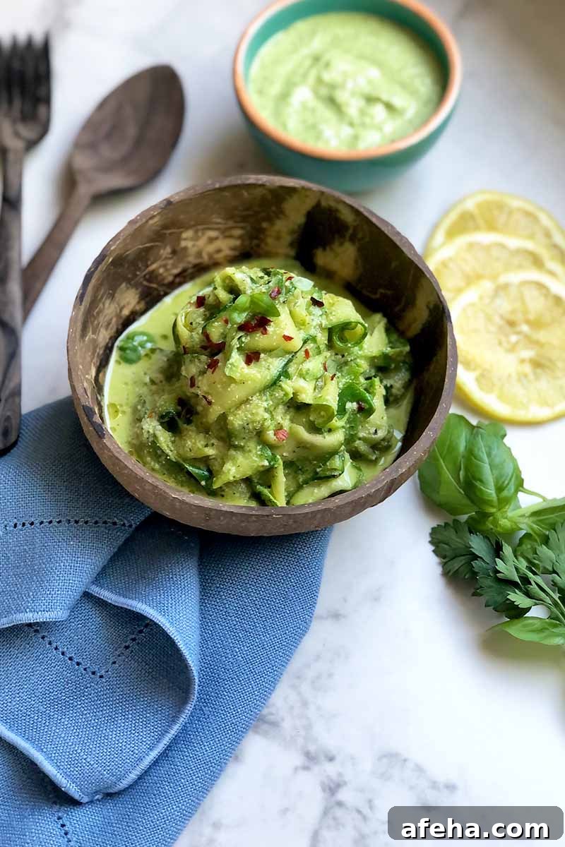Vertical image of a wooden bowl filled with a pile of cooked thinly sliced vegetables covered in a vibrant green sauce and fresh herb garnish next to a blue towel, wooden spoons, lemon slices, and a bowl filled with sauce.