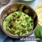 Horizontal image of a wooden bowl filled with a pile of cooked thinly sliced vegetables covered in a vibrant green sauce and fresh herb garnish next to a blue towel, wooden spoons, lemon slices.