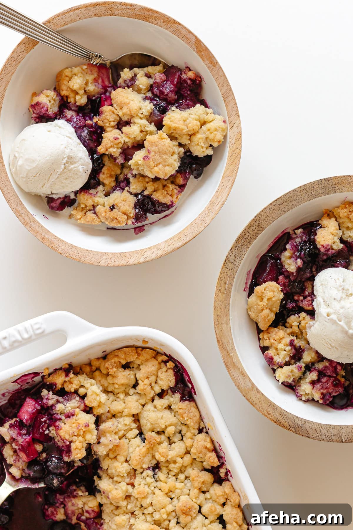 Two bowls of blueberry apple crumble topped with vanilla ice cream next to the baking dish.