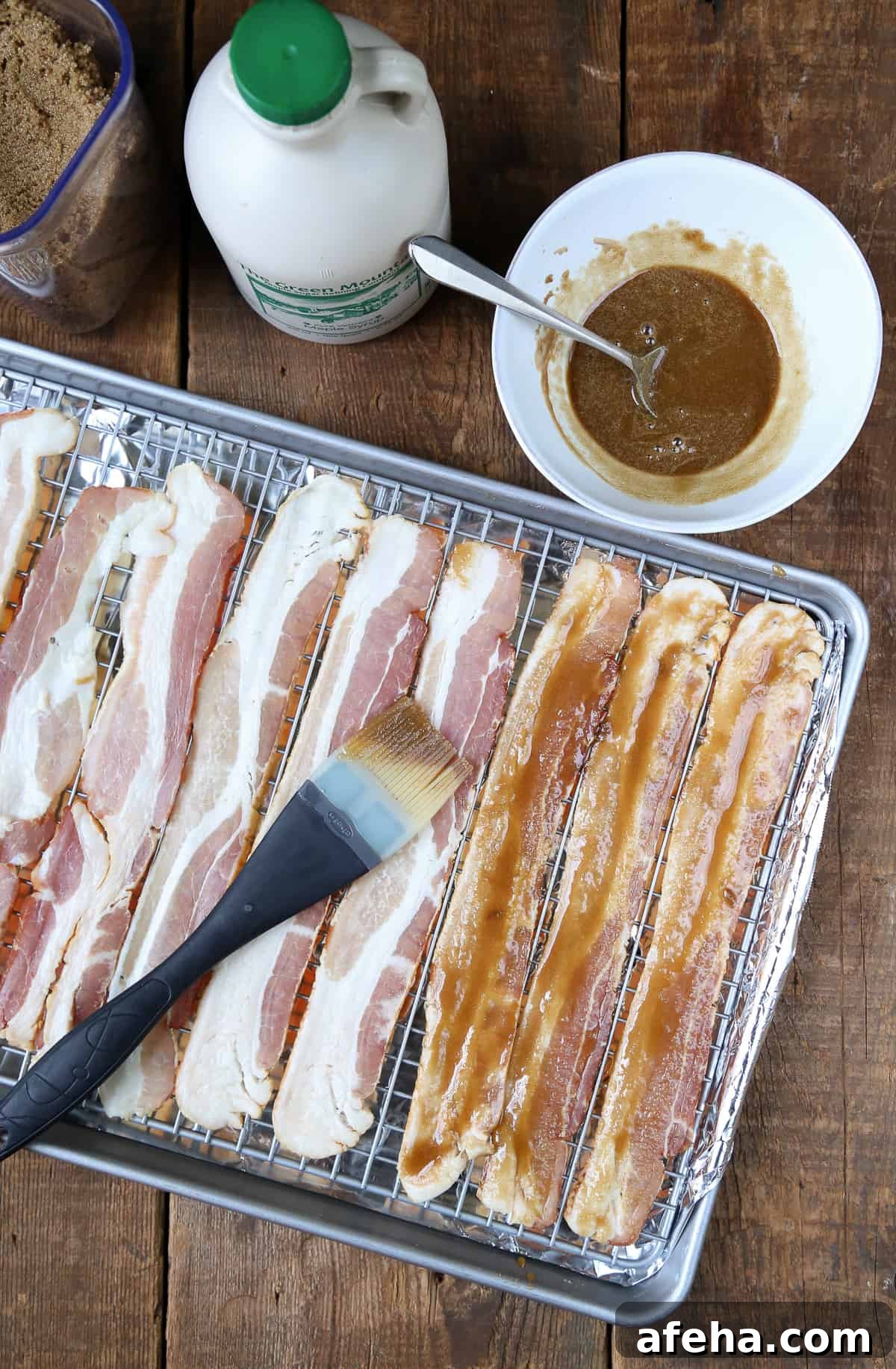 Praline-Crusted Candied Bacon — Sweet & Crunchy Bacon Recipe 5 uncooked bacon on a sheet pan being brushed with a brown sugar mixture.
