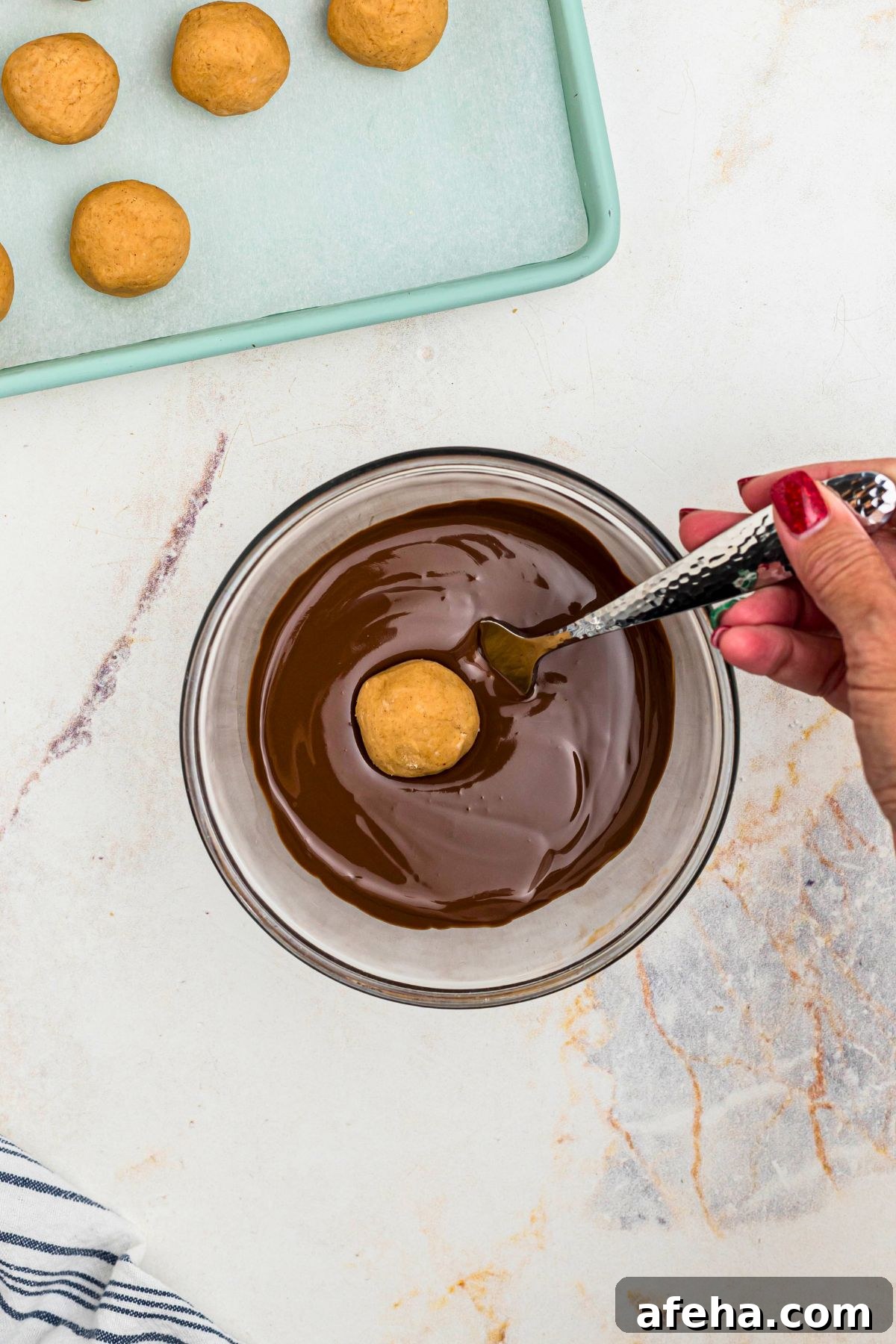 Melted chocolate in a clear glass bowl, with a peanut butter ball being dipped into the bowl. 