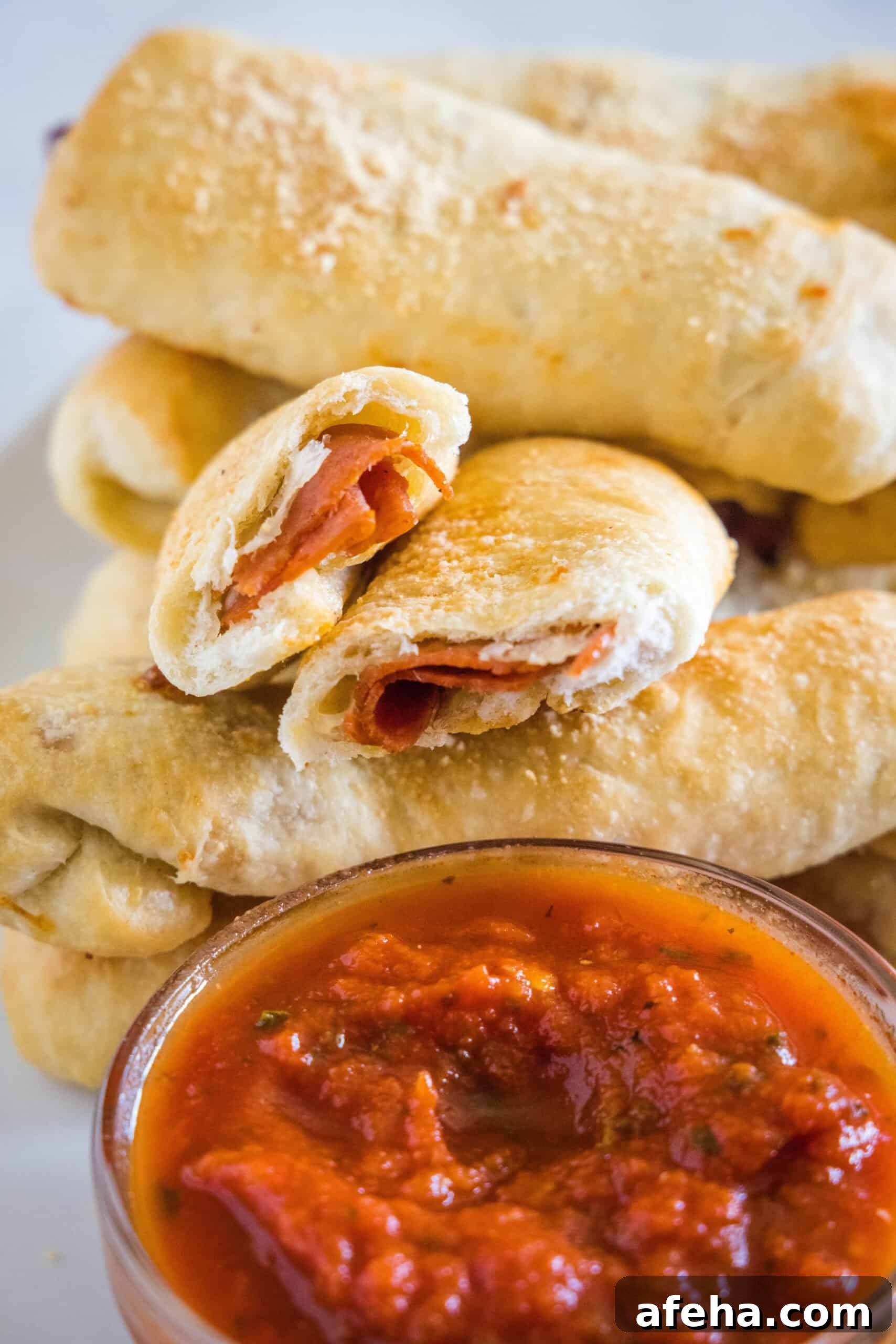 Pepperoni rolls stacked behind a bowl of marinara sauce, with one roll broken in half.