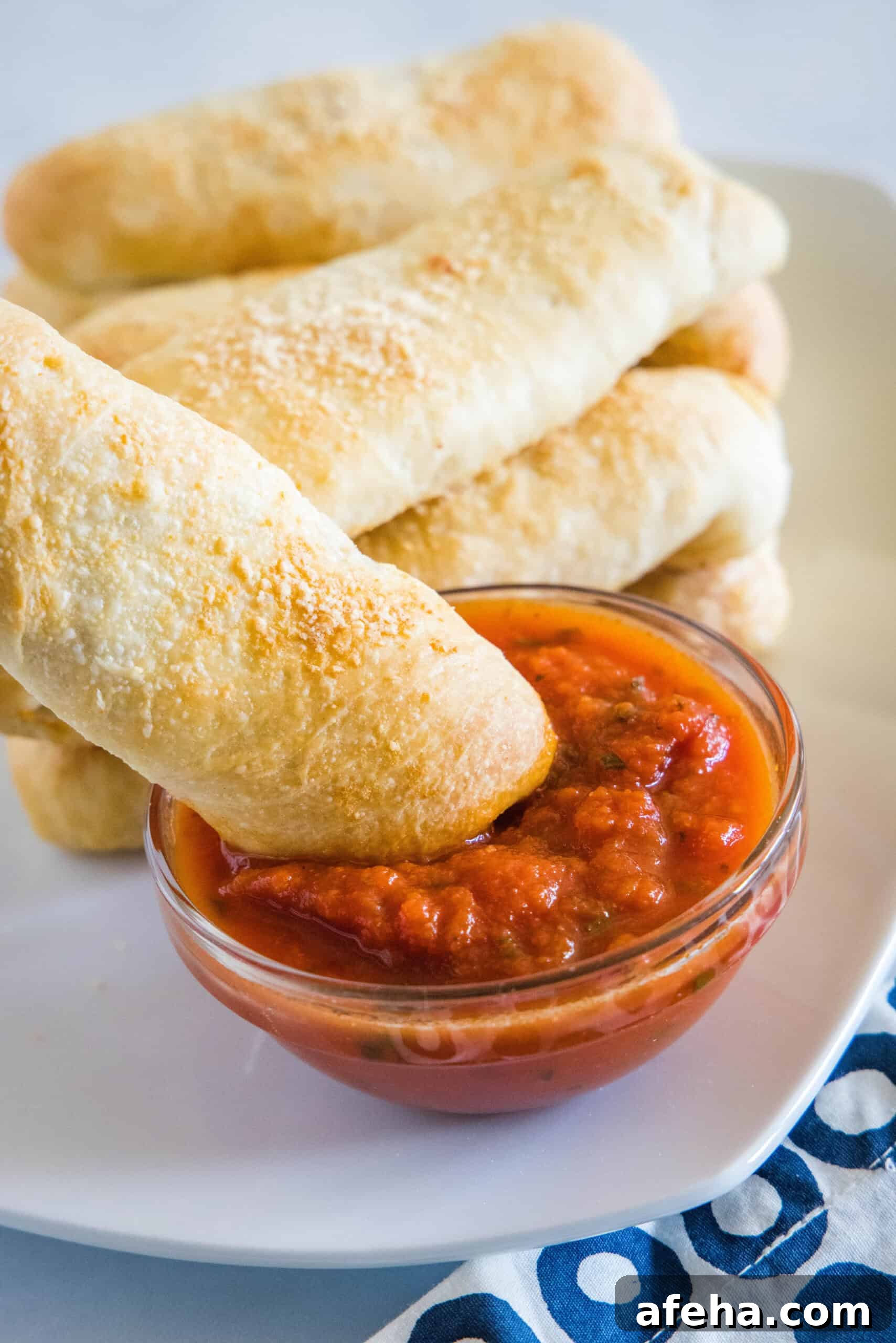 A pepperoni roll being dipped into a small bowl of marinara sauce with more rolls in the background.