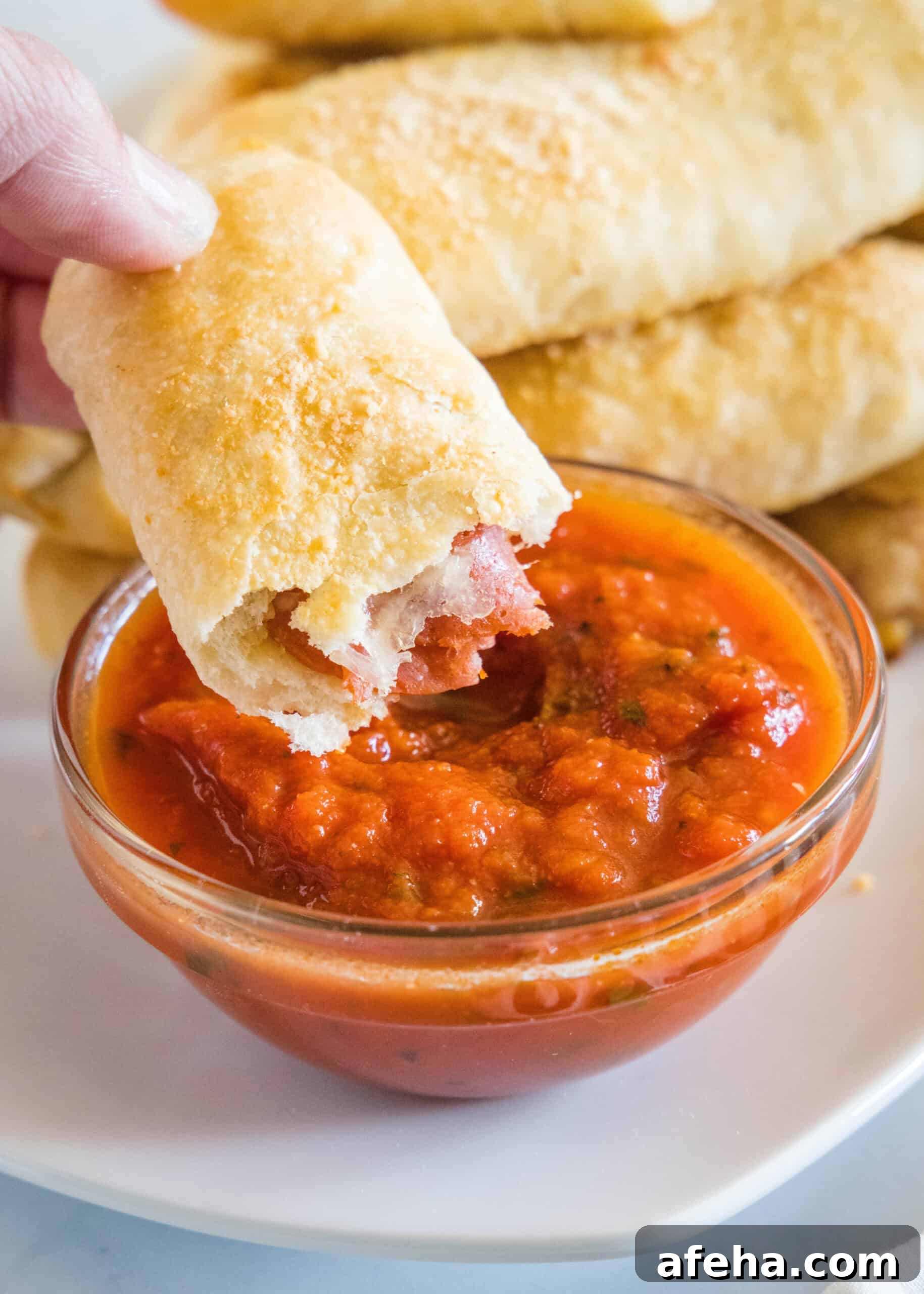 A hand holding one half of a pepperoni roll above a small bowl of marinara sauce with more rolls in the background.