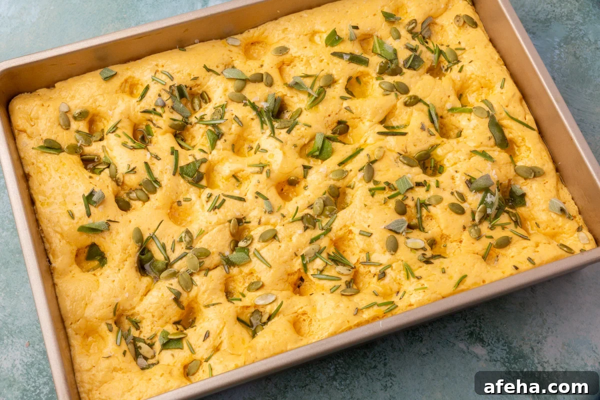 Unbaked pumpkin focaccia dough dimpled and topped with rosemary, sage, pepitas, and sea salt.
