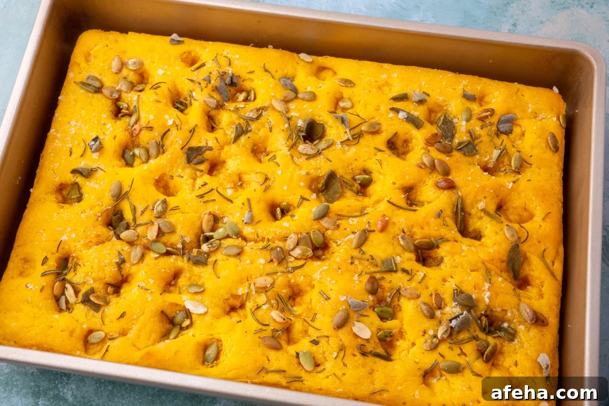 Freshly baked gluten-free pumpkin focaccia in a pan topped with rosemary, sage, pepitas, and flaky sea salt.