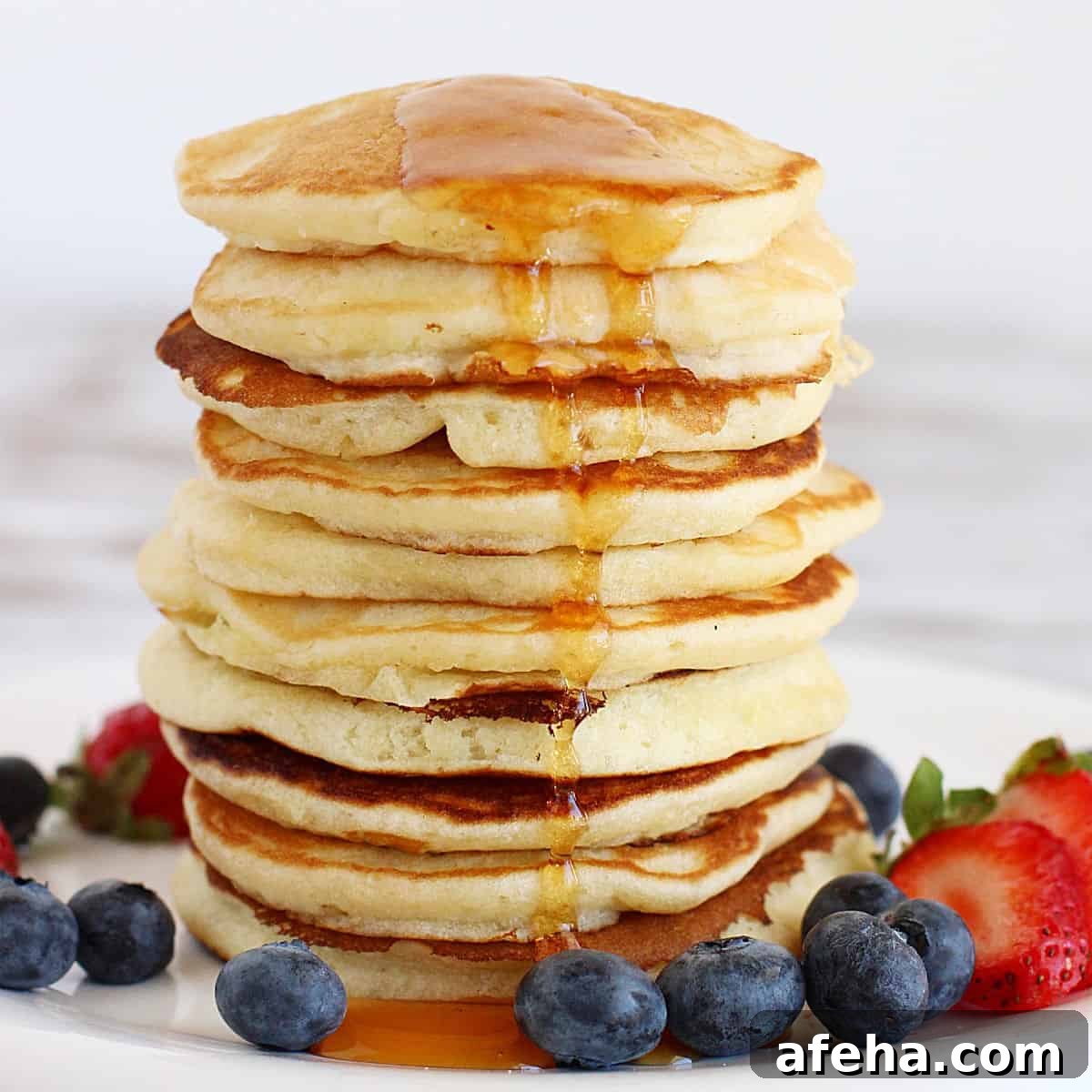 Tall stack of buttermilk pancakes with maple syrup and berries around. White background.