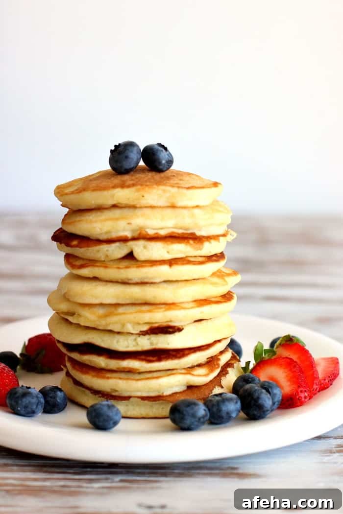 Berries top and around a stack of pancakes on a white plate, white table.
