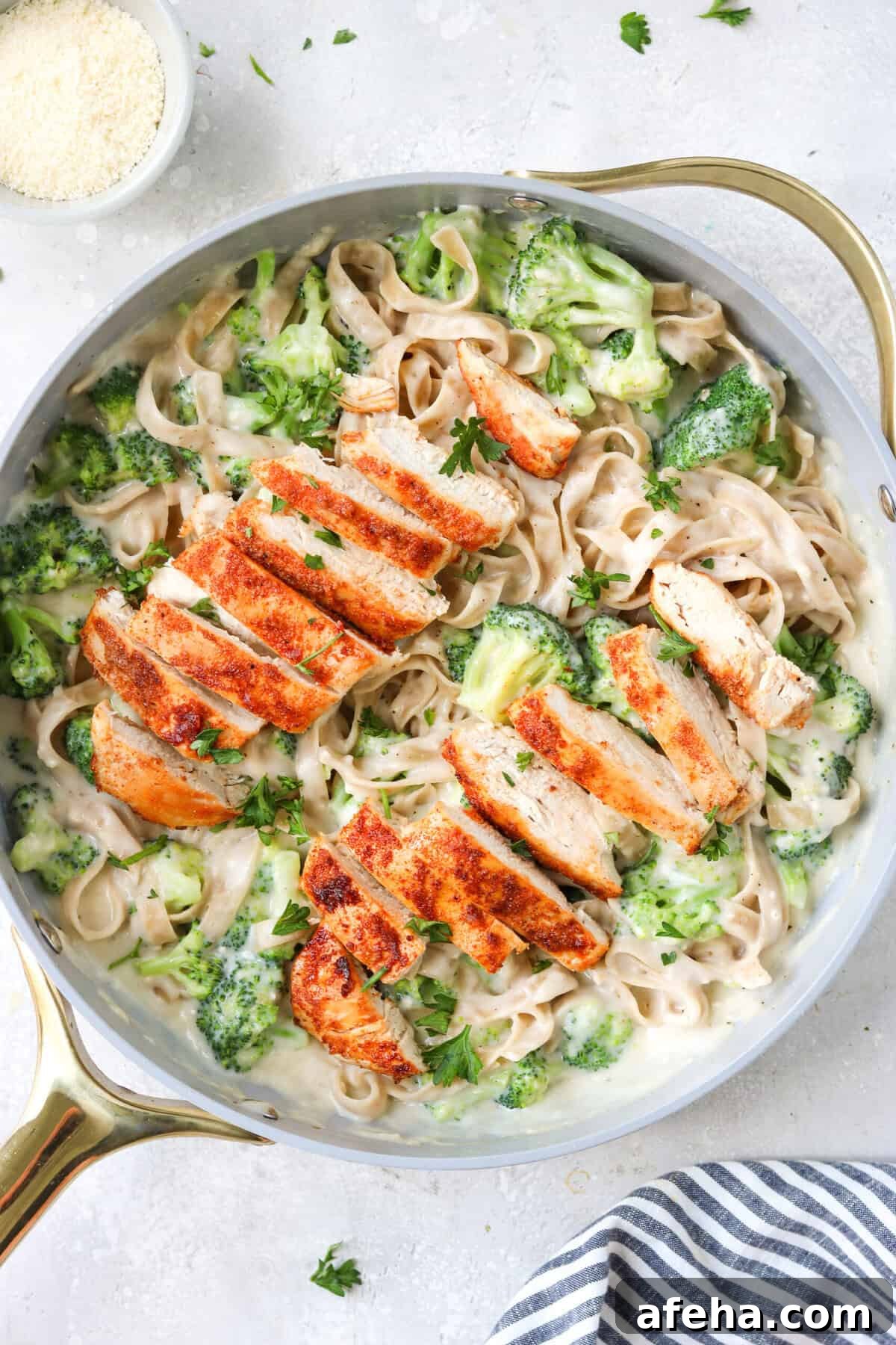 chicken and broccoli with alfredo sauce and gluten free pasta in a skillet with a napkin and a small bowl of parmesan on the side