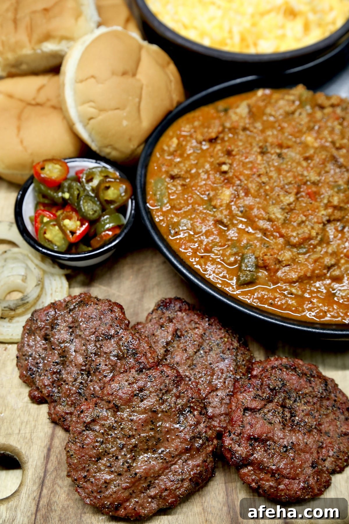 Grilled burger patties on a board with chili, cheese, jalapenos, buns.