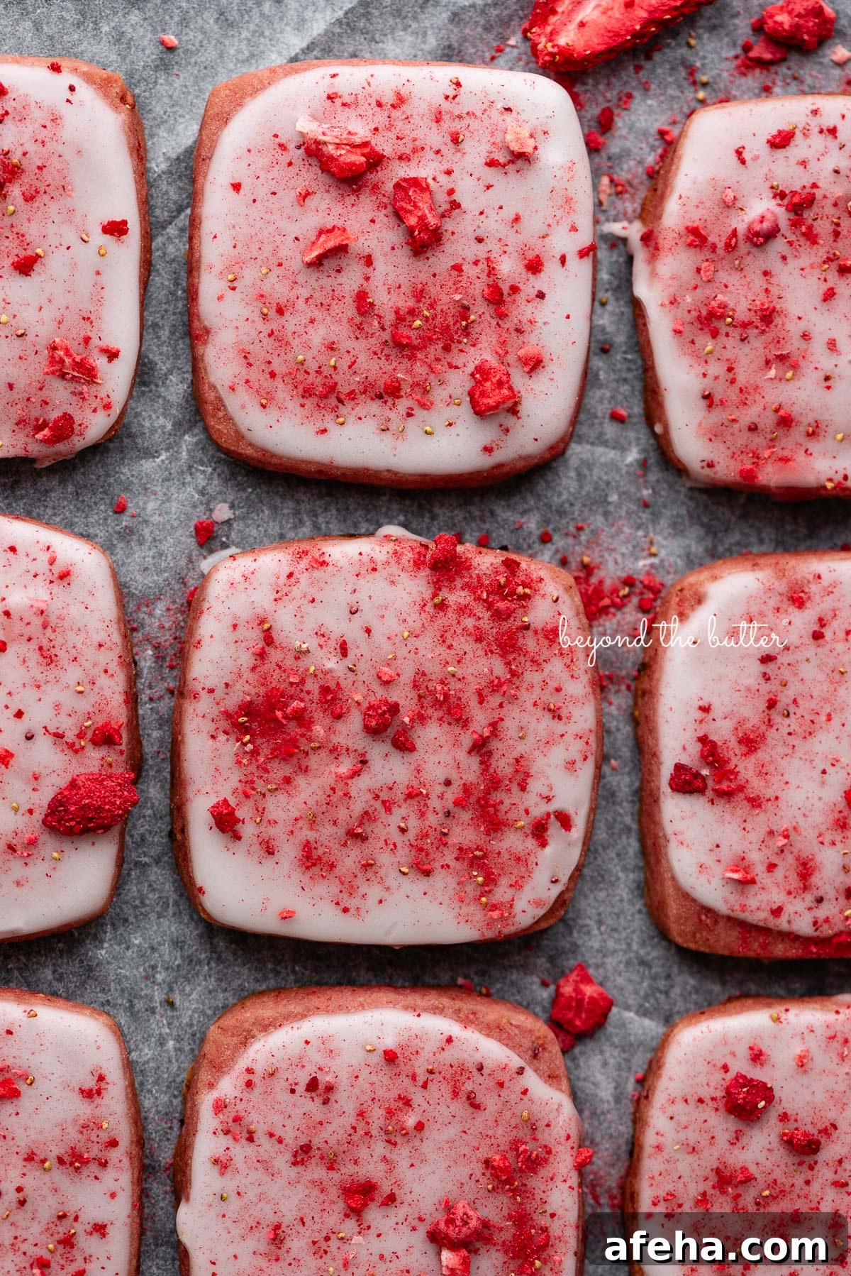 Rows of strawberry shortbread cookies topped with a powdered sugar glaze and freeze-dried strawberry bits on a parchment paper and black background.