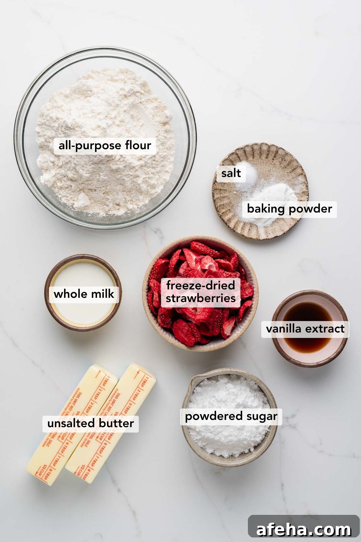 Ingredients in various size bowls and plates labeled as all purpose flour, salt, baking powder, whole milk, freeze dried strawberries, vanilla extract, unsalted butter, and powdered sugar on a white marbled background.