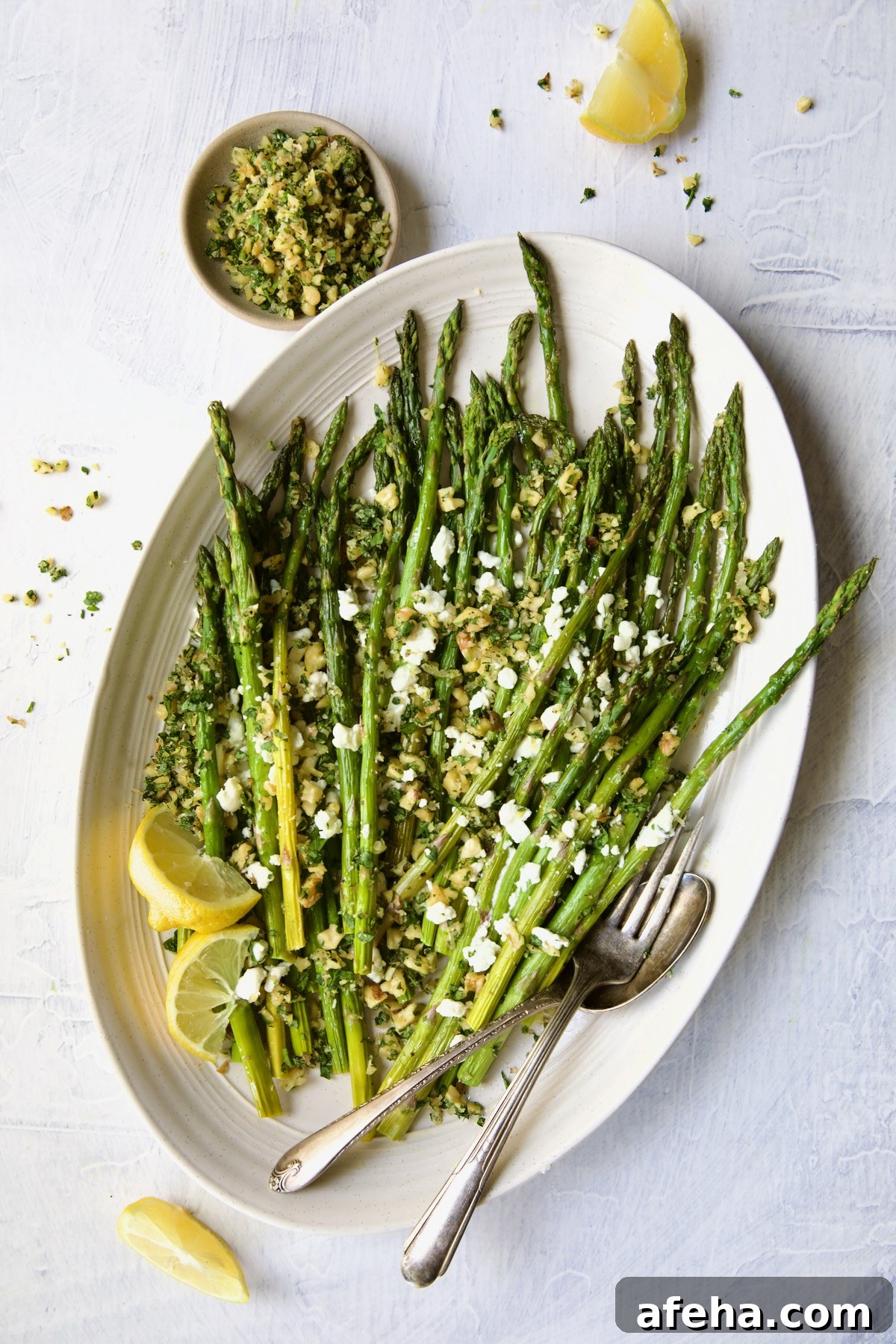 Roasted Asparagus with Walnut Lemon Gremolata and Goat Cheese on white oval platter on an angle with lemon wedges, serving utensils and bowl of gremolata.