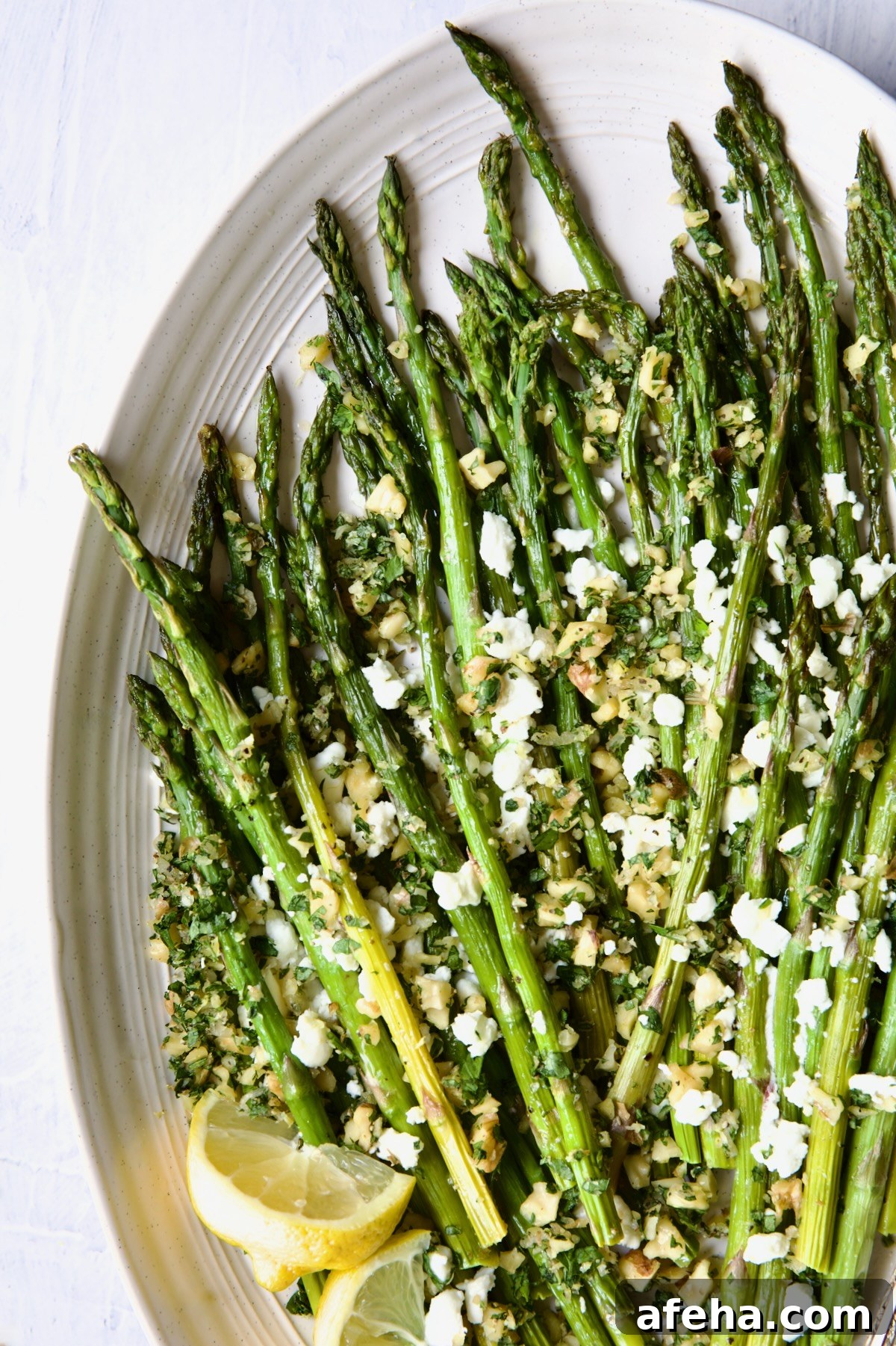 Roasted Asparagus with Walnut Lemon Gremolata and Goat Cheese on white oval platter with lemon wedges.